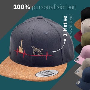 Op de afbeelding: Marineblauwe snapback cap met een kurken klep. De cap heeft een rood en bruin gitaar- en drumstel ontwerp met een hartslaglijn. De tekst "100% personalisierbar!" staat bovenaan. De cap maakt deel uit van een collectie met meerdere kleuropties.