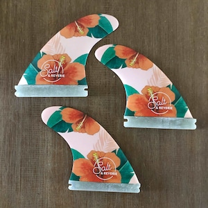 Op de afbeelding: Drie roze surfplankvinnen met een tropisch bloemenontwerp met hibiscusbloemen en groene bladeren. De vinnen hebben een witte basis met de tekst "Salt & Reverie" erop gedrukt.