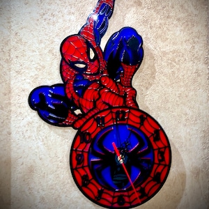 Pode incluir: Um relógio de parede vermelho e azul com um design de personagem do Homem-Aranha. O mostrador do relógio tem um design de teia de aranha preta com fundo azul e números vermelhos.