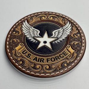 Puede incluir: Un posavasos redondo de cuero marrón con las palabras "U.S. AIR FORCE" en relieve en el borde. El centro presenta una estrella blanca, alas plateadas y un fondo negro. El posavasos tiene un diseño decorativo tallado.