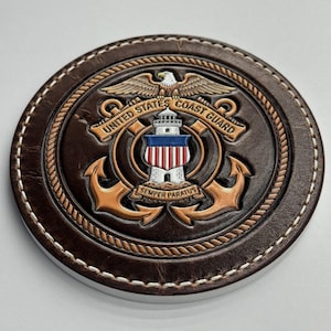 May include: A round, brown leather coaster with a stitched edge. The center features the United States Coast Guard emblem, including an eagle, lighthouse, anchors, and the text "United States Coast Guard" and "Semper Paratus".