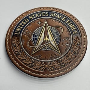 Puede incluir: Un objeto redondo de cuero marrón con las palabras "UNITED STATES SPACE FORCE" en letras doradas. El centro presenta un emblema estilizado con una estrella, un globo terráqueo y un cohete. El borde tiene un borde cosido y un diseño de hojas decorativas. Aproximadamente 10 cm de diámetro.