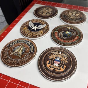 May include: A collection of brown leather coasters featuring embossed emblems of the United States military branches, including the Air Force, Space Force, Marine Corps, and Coast Guard. Each coaster has a white border and detailed stitching.