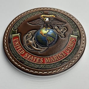 Puede incluir: Un objeto redondo de cuero marrón con el emblema de los Marines de los Estados Unidos. El emblema presenta un águila, un globo terráqueo y un ancla, con las palabras "United States Marine Corps" y "Semper Fi". Tiene un borde cosido.