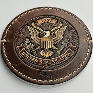 Puede incluir: Un posavasos redondo de cuero marrón con un diseño en relieve detallado. El centro presenta un emblema dorado del Ejército de los Estados Unidos con el texto "UNITED STATES ARMY" y "1775". El borde está cosido con hilo blanco.