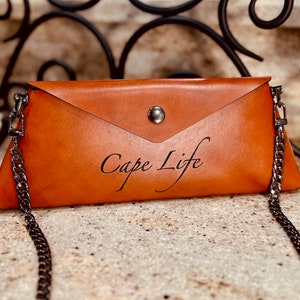 May include: A brown leather envelope clutch with a silver chain strap. The clutch has a silver button closure and the text "Cape Life" is embossed on the front.