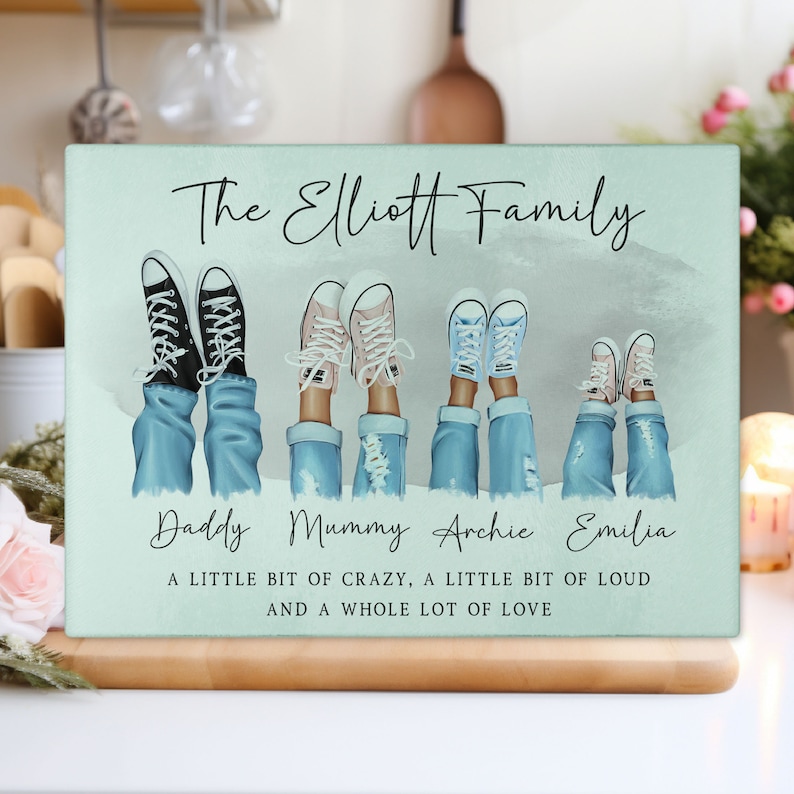 May include: A rectangular, pale green family-themed decorative sign. The sign features illustrations of legs in jeans and trainers, with the text "The Elliott Family" and names below. The bottom text reads "A LITTLE BIT OF CRAZY, A LITTLE BIT OF LOUD AND A WHOLE LOT OF LOVE."