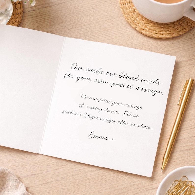 May include: An open white greeting card with handwritten text: "Our cards are blank inside for your own special message." A gold pen and small white bowl with paper clips are on the right. A cup of coffee sits in the background.