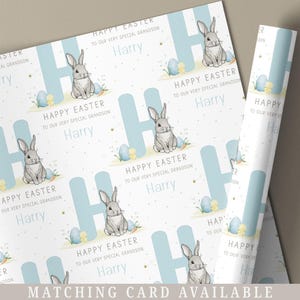 May include: White wrapping paper with a repeating pattern of blue letters "H" with a grey bunny sitting on each letter. The bunny is surrounded by yellow and blue Easter eggs. The text "Happy Easter to our very special grandson Harry" is repeated throughout the pattern.