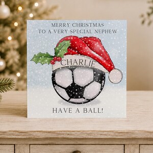 May include: A square Christmas greeting card featuring a soccer ball wearing a red Santa hat with green holly leaves and red berries. The text reads MERRY CHRISTMAS TO A VERY SPECIAL NEPHEW, CHARLIE, and HAVE A BALL! against a light blue snowy background.