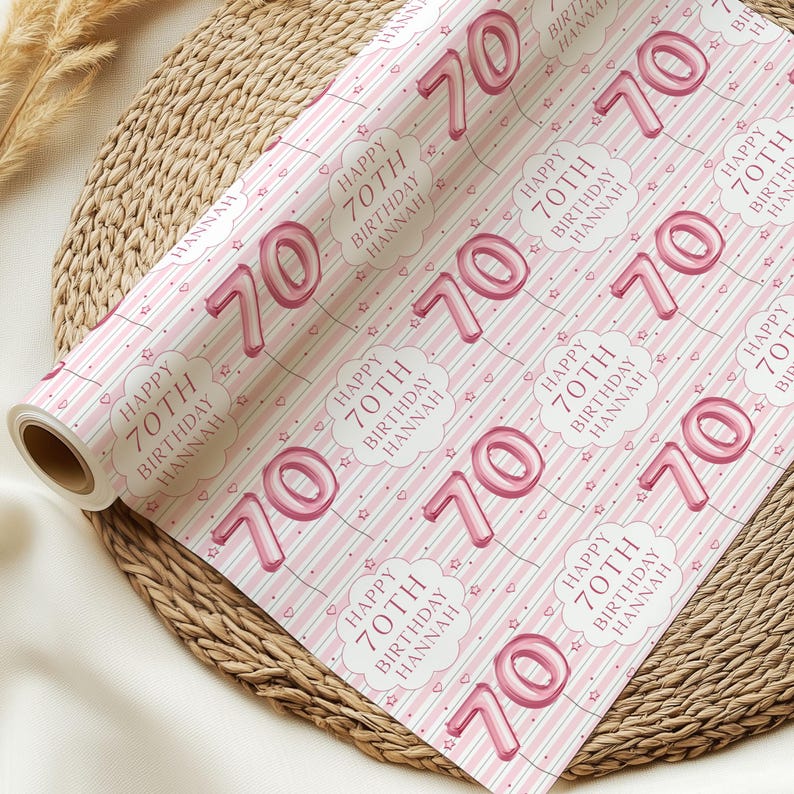May include: Pink wrapping paper roll with a repeating pattern of the number 70 in a balloon font, clouds, hearts, and stars. The text "HAPPY 70TH BIRTHDAY HANNAH" is also featured. The paper has pink and white stripes.