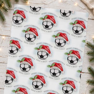 May include: Wrapping paper featuring a repeating pattern of soccer balls wearing red Santa hats with green holly leaves. Each ball has the name Lucas printed on a white band. The text Merry Christmas and Have a Ball is printed in black.