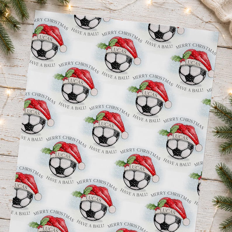 May include: Wrapping paper featuring a repeating pattern of soccer balls wearing red Santa hats with green holly leaves. Each ball has the name Lucas printed on a white band. The text Merry Christmas and Have a Ball is printed in black.