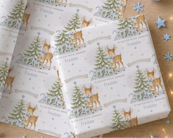May include: White wrapping paper featuring a repeating pattern of brown deer with blue bows, green Christmas trees, blue stars, and the text 2026, To our very special grandson, Freddie, and Happy 1st Christmas printed in a light blue and grey font.