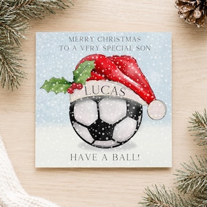 May include: A square greeting card featuring a soccer ball wearing a red Santa hat with green holly and red berries. The text reads MERRY CHRISTMAS TO A VERY SPECIAL SON, LUCAS, and HAVE A BALL! The background is a light blue snowy winter scene.