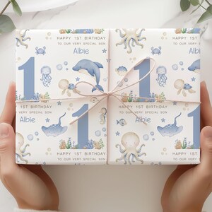 Puede incluir: Regalo envuelto con un dise&ntilde;o de tem&aacute;tica marina. El papel de regalo presenta ilustraciones de criaturas marinas, un gran n&uacute;mero 1 azul y el texto "HAPPY 1ST BIRTHDAY TO OUR VERY SPECIAL SON Albie". Atado con hilo.