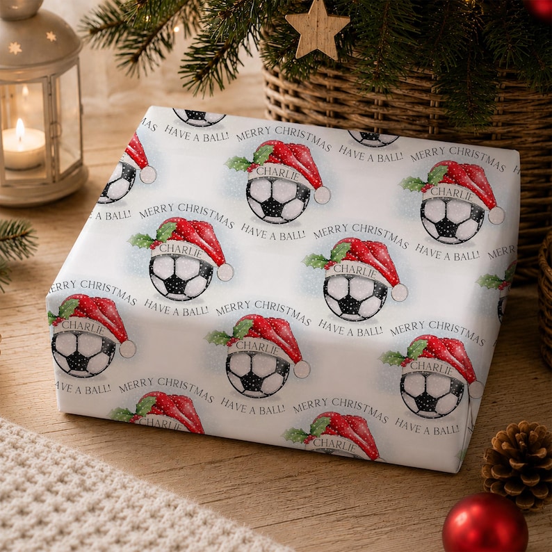 May include: White wrapping paper featuring a repeating pattern of soccer balls wearing red Santa hats with green holly leaves. Each hat includes the name Charlie printed in black text. The text Merry Christmas and Have a Ball is printed.
