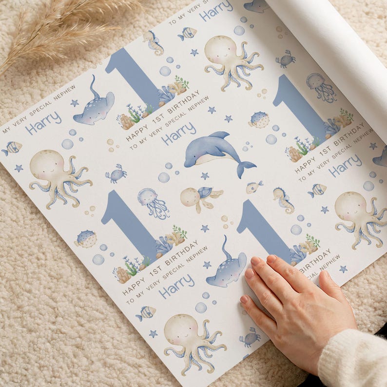 Puede incluir: Papel de regalo blanco con un dise&ntilde;o marino azul y acuarela. El dise&ntilde;o incluye pulpos, delfines y el n&uacute;mero 1. El texto dice "Happy 1st Birthday" y "Harry".