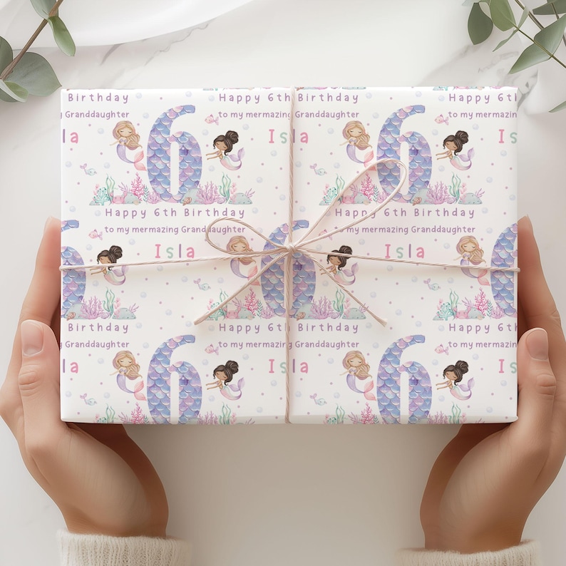 May include: White gift wrap with a mermaid theme. The wrap features a repeating pattern of mermaids, seashells, and the number six. The text "Happy 6th Birthday to my mermazing Granddaughter Isla" is repeated throughout the wrap.