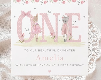 Puede incluir: Tarjeta rosa y blanca con la palabra "ONE" en letras grandes de color rosa con lunares blancos. Las letras están decoradas con un ciervo y un gato que llevan vestidos rosas. La tarjeta dice "To our beautiful daughter Amelia with lots of love on your first birthday."