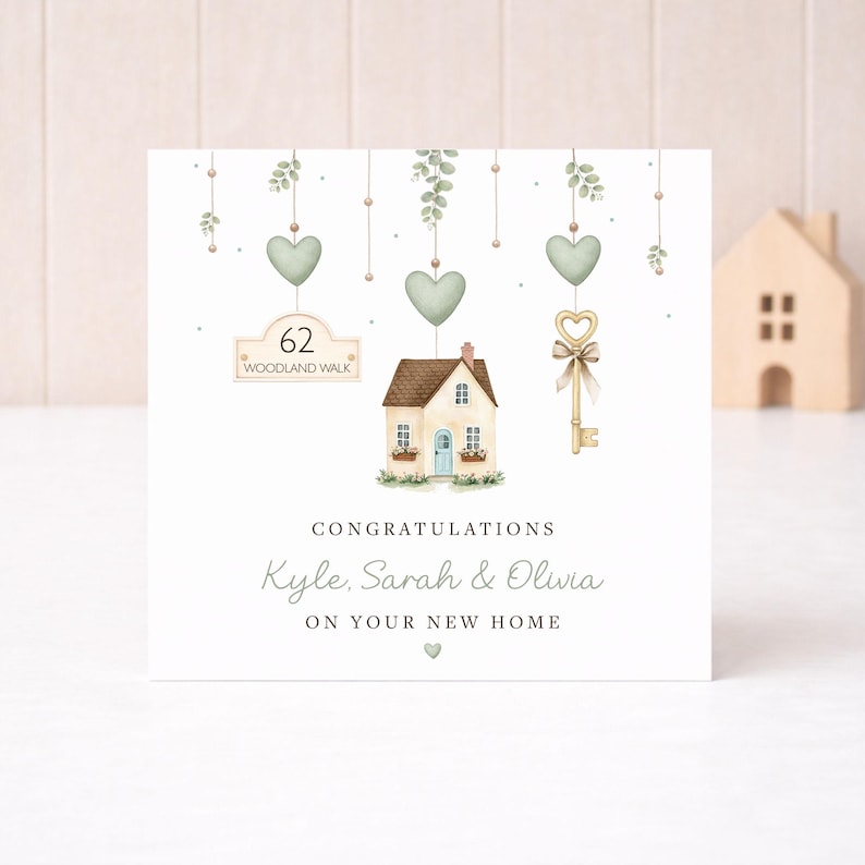 May include: A new home greeting card with a watercolour illustration. The card features a small house, a key, and hanging hearts. The text reads "Congratulations Kyle, Sarah & Olivia on your new home" and "62 Woodland Walk".