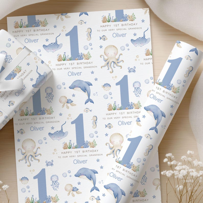 Puede incluir: Papel de regalo con tem&aacute;tica marina, con un n&uacute;mero uno azul, delfines, pulpos y otras criaturas marinas. El texto dice "Happy 1st Birthday" y "Oliver". Tambi&eacute;n se ve un rollo de papel a juego.