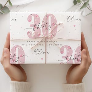 May include: Pink floral patterned gift wrap with the number 30 in pink with the word "thirty" written in black script. The text "Happy 30th Birthday to a very special daughter Olivia" is printed on the wrapping paper.