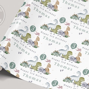 May include: White wrapping paper with a repeating pattern of green, blue and pink dinosaurs. The text "Happy 3rd Birthday Thomas" is repeated throughout the pattern. The number "3" is also repeated throughout the pattern.