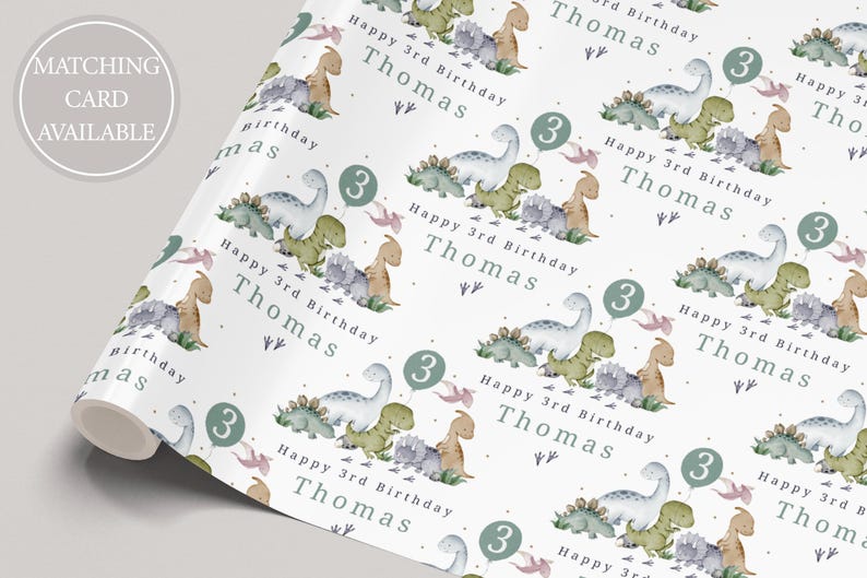 May include: White wrapping paper with a repeating pattern of green, blue and pink dinosaurs. The text "Happy 3rd Birthday Thomas" is repeated throughout the pattern. The number "3" is also repeated throughout the pattern.