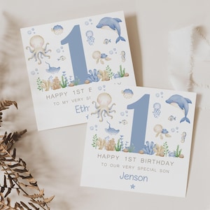 Puede incluir: Dos tarjetas de cumplea&ntilde;os cuadradas con un tema oce&aacute;nico de acuarela. Cada tarjeta presenta un gran n&uacute;mero 1 azul, un delf&iacute;n, un pulpo y otras criaturas marinas. El texto dice "Feliz 1er cumplea&ntilde;os" y un nombre.