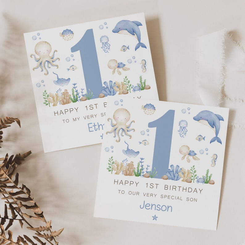 Puede incluir: Dos tarjetas de cumplea&ntilde;os cuadradas con un tema oce&aacute;nico de acuarela. Cada tarjeta presenta un gran n&uacute;mero 1 azul, un delf&iacute;n, un pulpo y otras criaturas marinas. El texto dice "Feliz 1er cumplea&ntilde;os" y un nombre.