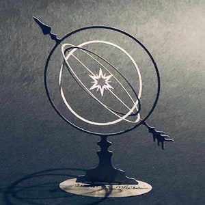 Puede incluir: Una escultura de metal negro de una esfera celeste con una estrella en el centro. La esfera está rodeada por dos anillos, y la escultura está montada sobre una base con una flecha apuntando hacia la derecha.