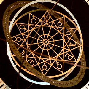 Op de afbeelding: Een close-up van een decoratieve wijzerplaat met een ingewikkeld, goudkleurig ontwerp met een stervormig patroon en astrologische symbolen. De wijzerplaat is omgeven door een zwarte achtergrond.