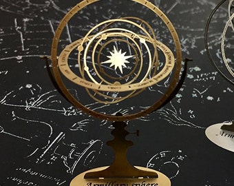 Op de afbeelding: Een goudkleurige armillairsfeer met een stervormig middelpunt en concentrische ringen, gemonteerd op een standaard. De basis leest "Armillary sphere". De achtergrond is een donkere kaart.