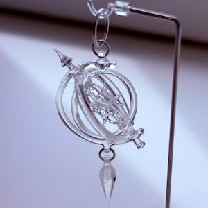 May include: A clear, transparent glass pendant with a complex, ornate design. The pendant is suspended from a metal stand, showcasing its intricate details. The design includes circular elements and a pointed bottom, creating a unique, artistic piece.