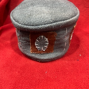 May include: A small, round, gray fabric container with a lid. The container has brown patches with white stitched designs. The item is sitting on a red surface. The container appears to be handmade.