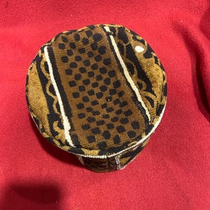 May include: A round, patterned hat with a brown and black spotted design. The hat has a white trim and is set against a red background. The hat's design includes black and white stripes.