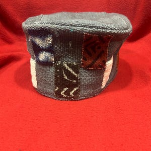 May include: A handcrafted, blue fabric hat with a cylindrical shape. The hat features decorative patches in various colors and patterns, including white, black, and red. The hat is set against a red background.