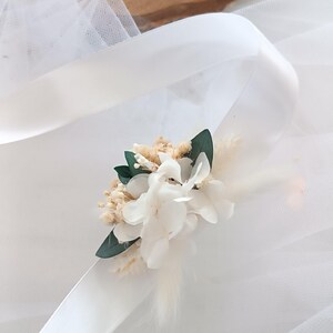 May include: A white satin ribbon with a small bouquet of dried white flowers and green leaves attached. The bouquet is tied with a white ribbon.