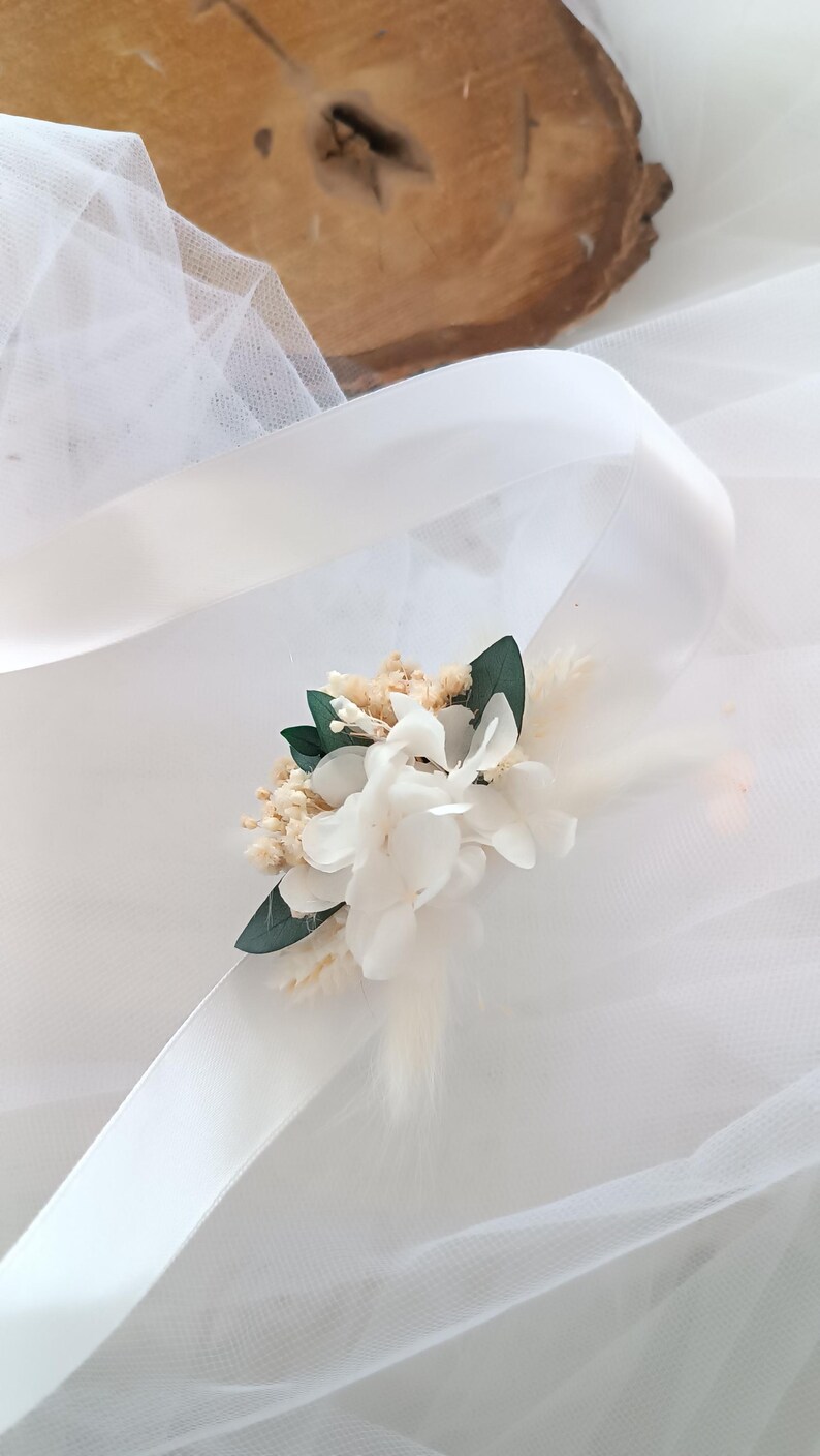 May include: A white satin ribbon with a small bouquet of dried white flowers and green leaves attached. The bouquet is tied with a white ribbon.