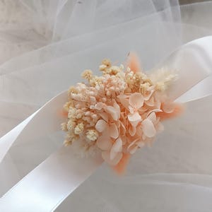 May include: A delicate bouquet of dried flowers in shades of peach and white, tied with a white satin ribbon. The flowers are arranged on a white tulle background.