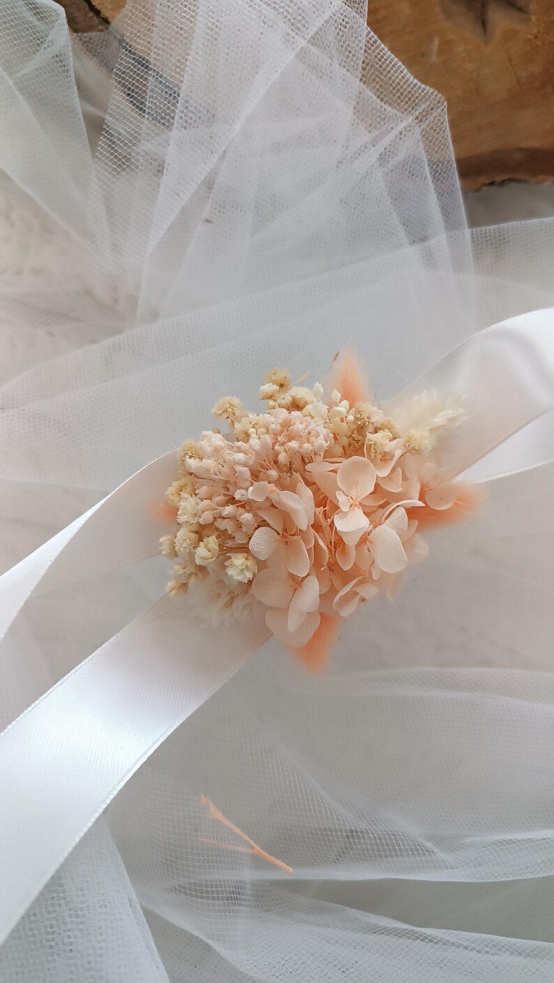May include: A delicate bouquet of dried flowers in shades of peach and white, tied with a white satin ribbon. The flowers are arranged on a white tulle background.
