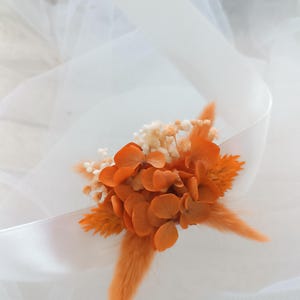 May include: A small bouquet of dried flowers in shades of orange and white, with a long, fluffy orange stem. The flowers are arranged on a white tulle fabric.