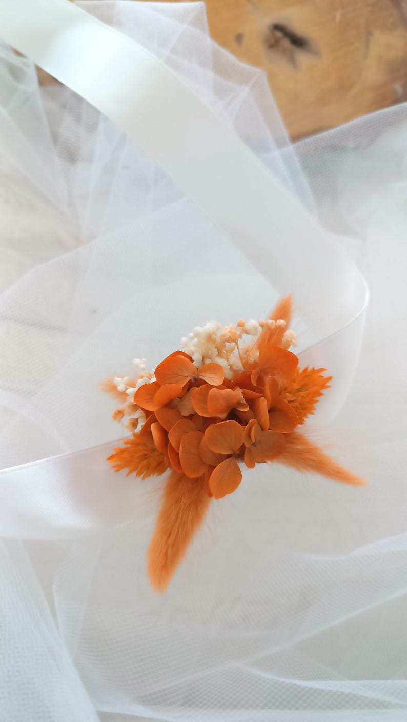 May include: A small bouquet of dried flowers in shades of orange and white, with a long, fluffy orange stem. The flowers are arranged on a white tulle fabric.
