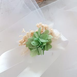 May include: A white satin ribbon with a green and white floral embellishment. The embellishment is made of dried flowers and greenery.