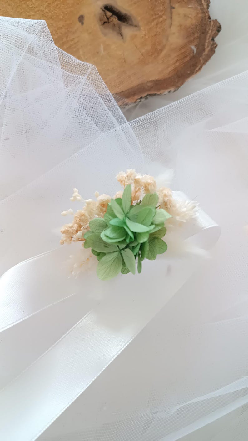 May include: A white satin ribbon with a green and white floral embellishment. The embellishment is made of dried flowers and greenery.