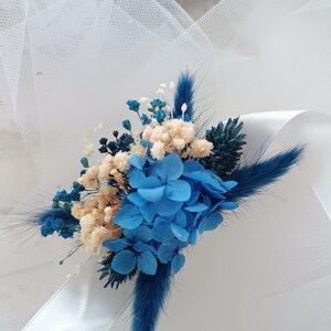 May include: A blue and white dried flower bouquet with a white satin ribbon. The bouquet features blue hydrangeas, white baby's breath, and blue dried grasses.