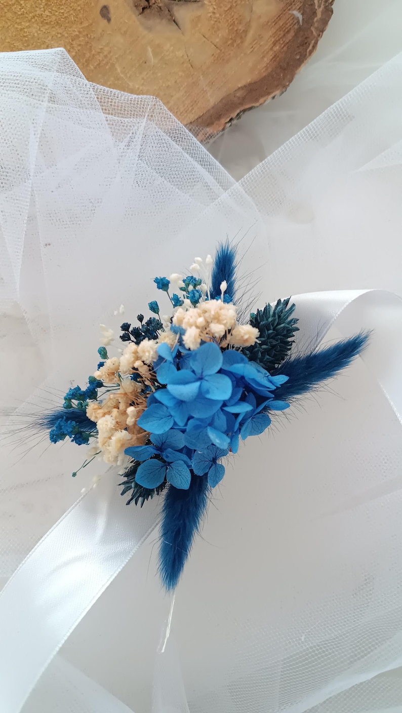 May include: A blue and white dried flower bouquet with a white satin ribbon. The bouquet features blue hydrangeas, white baby's breath, and blue dried grasses.