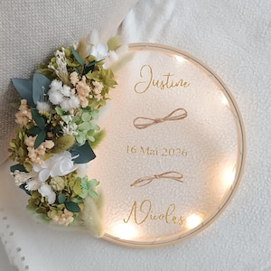Może przedstawiać: Okrągła, podświetlana dekoracja ścienna z imionami "Justine" i "Nicolas" w złotym skrypcie. Data "16 Mai 2026" jest również wyświetlana. Ozdobiona suszonymi kwiatami w kolorze białym, zielonym i beżowym.