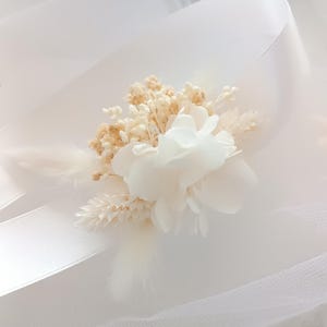 May include: A white flower bouquet with dried flowers and a white ribbon. The bouquet is on a white tulle background.